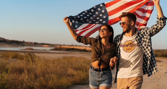 A couple in the outdoors with a man wearing a White Classic Logo Tee, both holding an end of the American Flag over their head as the two walk a dirt path.