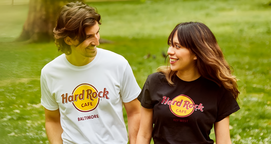 A couple walking in a meadow. A man wearing a white classic logo tee with the Hard Rock Cafe Baltimore logo with a woman wearing a black Classic Logo Tee with the Hard Rock Cafe Key West logo. The couple are staring at each other with a smile on their faces. 