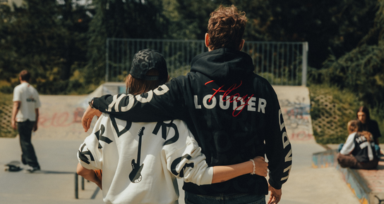 A woman wearing a cream white long sleeve sweater and a man wearing a black hoodie, both standing at a skate park.