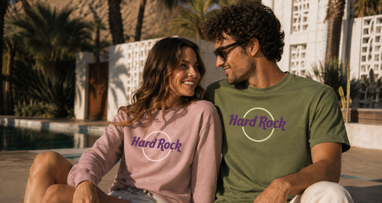 Two people wearing pastel Hard Rock apparel sit poolside at a sunny desert resort, showcasing the Color Vibes collection.
