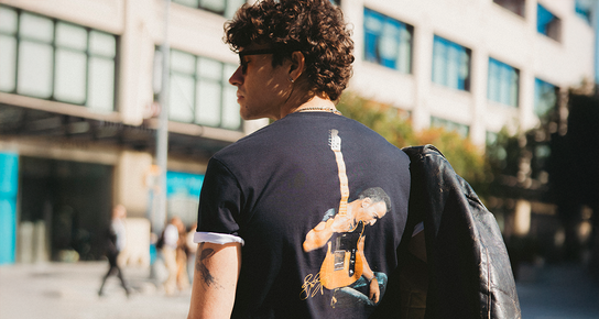 A person in an outdoor setting wearing a Hard Rock Bruce Springsteen Photograph T-shirt while wearing sunglasses and holding a backpack on one shoulder, looking to his left.  A person in an outdoor setting wearing a Hard Rock Bruce Springsteen Photograph T-shirt while wearing sunglasses and holding a backpack on one shoulder, looking to his left.