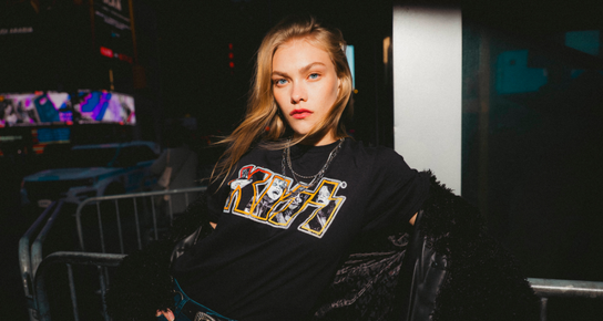 A woman in an indoor setting wearing a Hard Rock KISS tribute T-shirt while wearing a necklace, leaning on a short fence, looking to her left. 