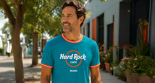 A couple walking in a meadow. A man wearing a white classic logo tee with the Hard Rock Cafe Baltimore logo with a woman wearing a black Classic Logo Tee with the Hard Rock Cafe Key West logo. The couple are staring at each other with a smile on their faces. 