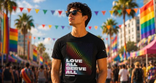 Man wearing a black Hard Rock Pride graphic tee standing at a colorful Pride festival with rainbow flags and palm trees in the background.